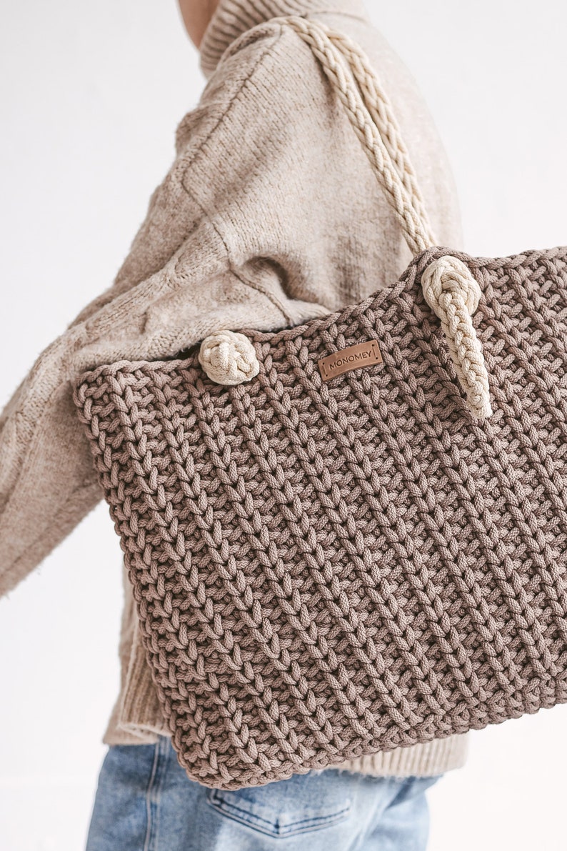 Op de afbeelding: Een bruine gehaakte tote bag met cr&egrave;mekleurige banden en een bruin leren label met de tekst "MONOMEY".