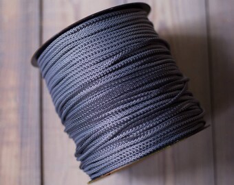 May include: A spool of gray rope, tightly wound and ready for use. The rope has a textured, woven appearance, and the spool is set against a light-colored wooden background. Ideal for crafting or general use.