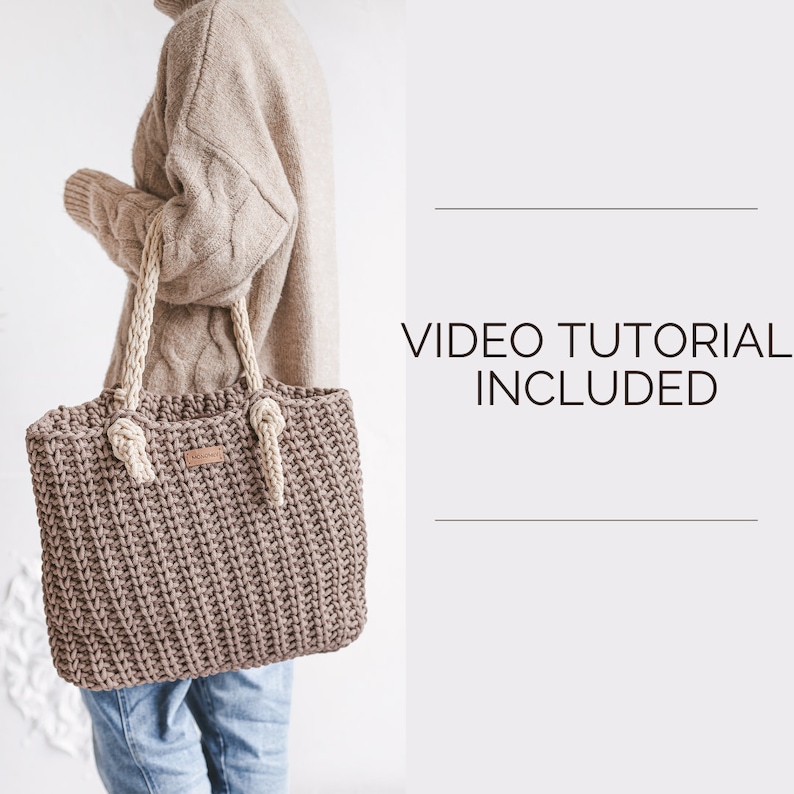 Op de afbeelding: Een bruine gehaakte tote bag met cr&egrave;mekleurige touwhandvatten. De tas wordt gedragen door iemand die een beige trui en een blauwe spijkerbroek draagt. De tekst "VIDEO TUTORIAL INCLUDED" wordt weergegeven op een witte achtergrond.