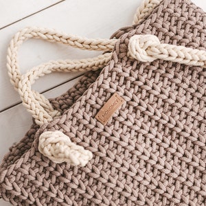 Op de afbeelding: Een bruine gehaakte tote bag met een cr&egrave;mekleurige gevlochten handvat en een bruin lederen label met de tekst "MONOMEY".