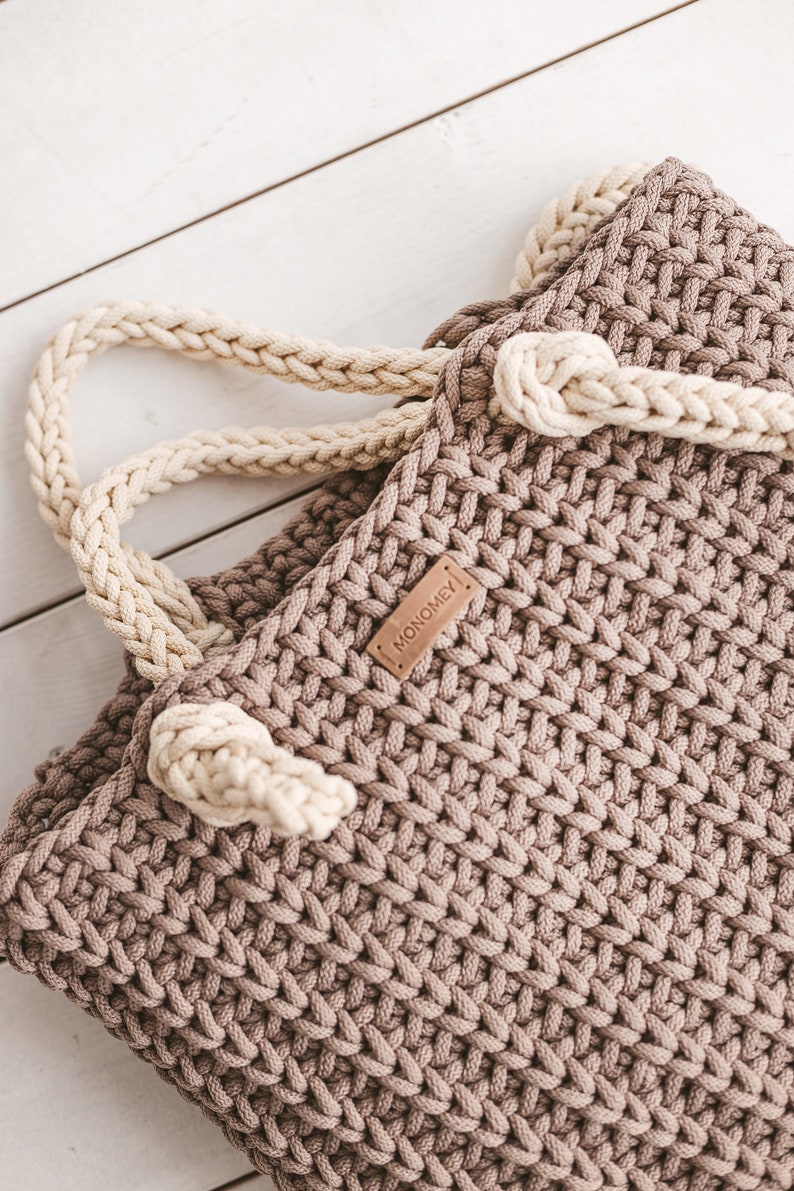 Op de afbeelding: Een bruine gehaakte tote bag met een cr&egrave;mekleurige gevlochten handvat en een bruin lederen label met de tekst "MONOMEY".