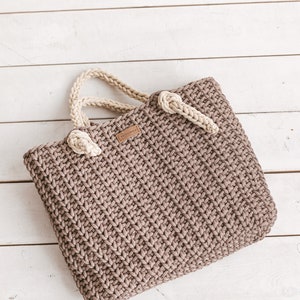 Op de afbeelding: Een bruine gehaakte tote bag met cr&egrave;mekleurige handvatten. De tas heeft een klein bruin leren label met het woord "HOMEMADE" erop.