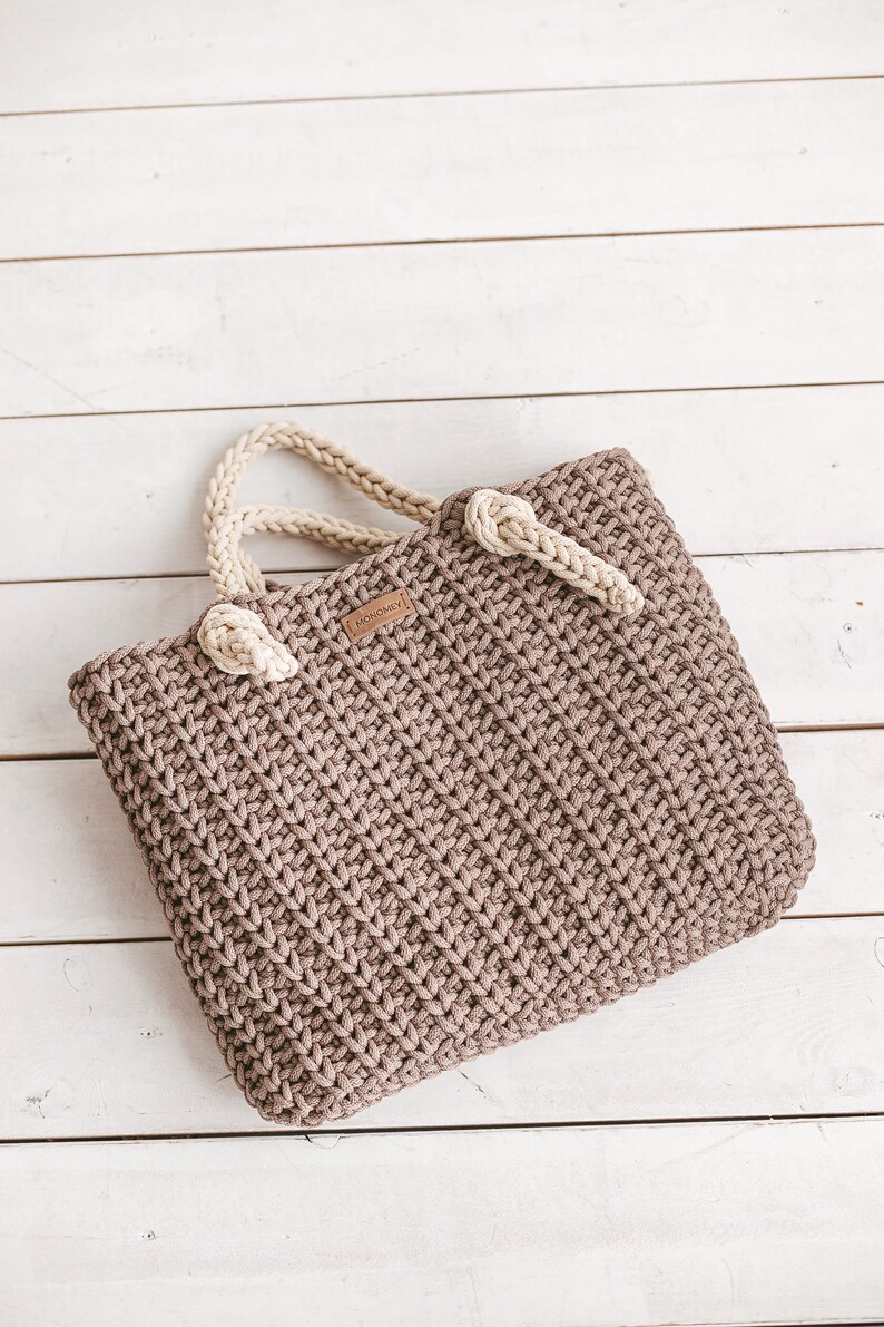 Op de afbeelding: Een bruine gehaakte tote bag met cr&egrave;mekleurige handvatten. De tas heeft een klein bruin leren label met het woord "HOMEMADE" erop.