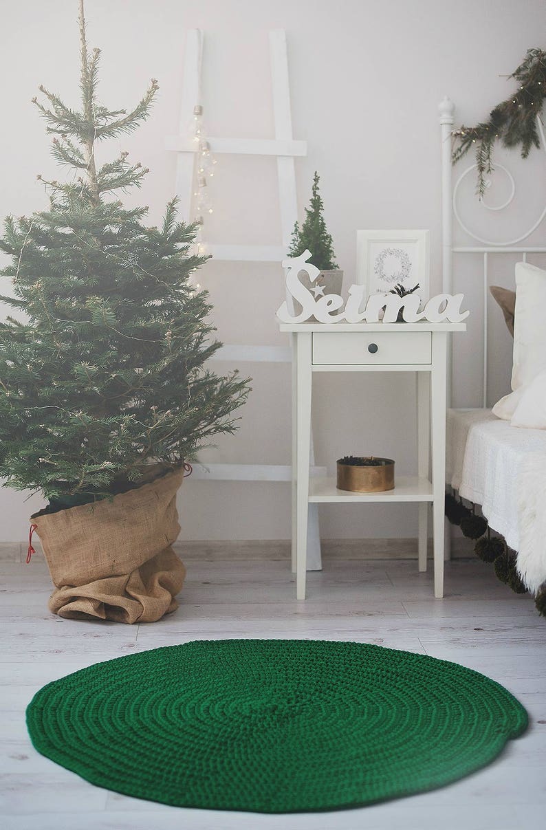 May include: A green crocheted round rug with a white wooden side table with a drawer and a shelf. The table has a white wooden sign that says "Selima" on it. There is a small potted Christmas tree on the table.