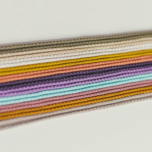 May include: A collection of colorful cords in various shades, including green, yellow, purple, and pink. The cords are tightly woven and arranged in parallel lines, creating a textured and visually appealing display. The cords are likely used for crafting or jewelry making.