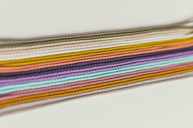 May include: A collection of colorful cords in various shades, including green, yellow, purple, and pink. The cords are tightly woven and arranged in parallel lines, creating a textured and visually appealing display. The cords are likely used for crafting or jewelry making.