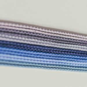 May include: Close-up of a bundle of blue, purple, and gray cords. The cords are tightly woven together, creating a textured surface. The color gradient transitions from light blue to dark blue and purple.