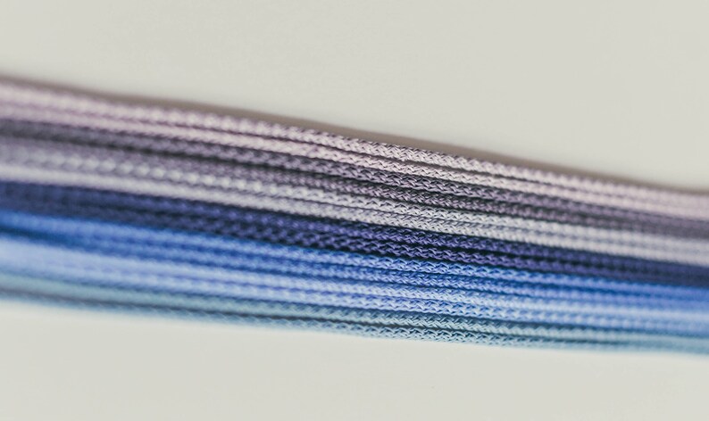 May include: Close-up of a bundle of blue, purple, and gray cords. The cords are tightly woven together, creating a textured surface. The color gradient transitions from light blue to dark blue and purple.