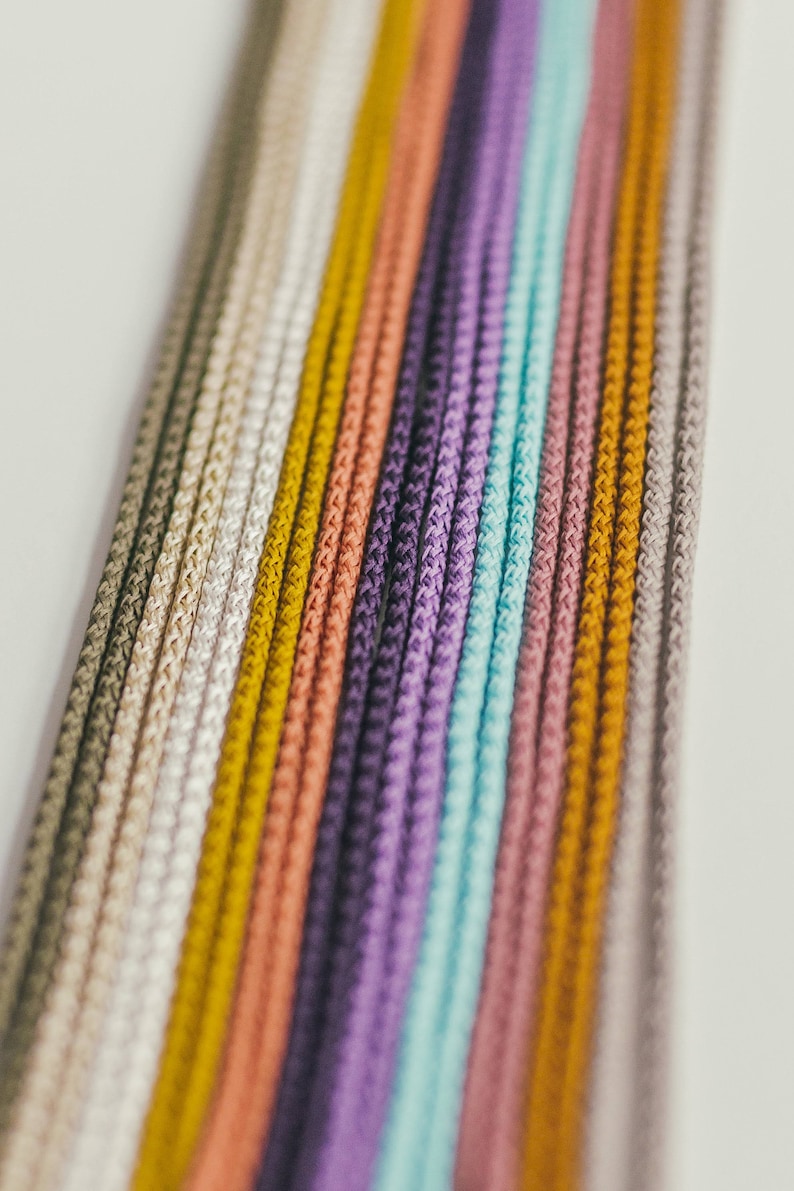 May include: Close-up of a collection of colorful cords in a variety of shades. The cords are arranged vertically, showcasing a spectrum of colors including beige, yellow, peach, purple, and teal. The cords have a textured, woven appearance.