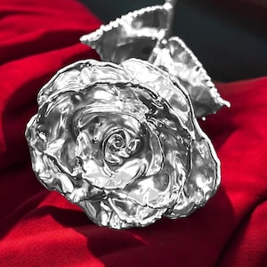 Op de afbeelding: Een enkele zilveren roos met een glanzende, metalen afwerking. De roos ligt op een rood fluwelen oppervlak.
