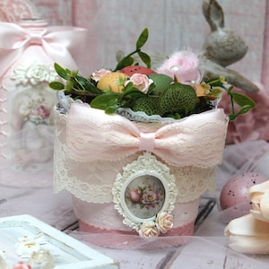 May include: A pink ceramic planter with a lace trim and a pink satin bow. The planter is filled with green plants, faux eggs, and a small framed floral print.