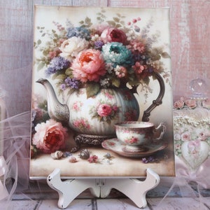 May include: A vintage-style painting of a teapot and teacup with a bouquet of pink, purple, and blue flowers. The teapot and teacup are decorated with floral patterns. The painting is on a white canvas with a white easel.