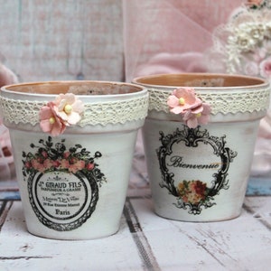 May include: Two white ceramic flower pots with lace trim and floral designs. One pot says "Bienvenue" and the other says "J. Giraud Fils Parfumeur a Grasse Maison De Luxe 20 Rue Etienne Marcel Paris".