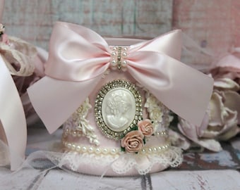 May include: A pink ceramic container with a cameo design, embellished with pearls, rhinestones, and a pink satin bow. The container is decorated with lace and faux flowers.