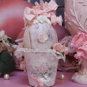 May include: A decorative egg-shaped container with a floral design, adorned with a pink bow and lace trim. The container is set in a matching lace-trimmed cup, with pearls scattered around. The overall theme is vintage and romantic.