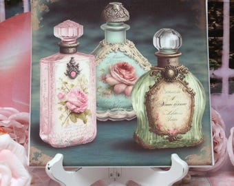 May include: A decorative print featuring three vintage perfume bottles. The bottles are in shades of pink, green, and blue, with ornate details and floral designs. The print is displayed on a white easel.