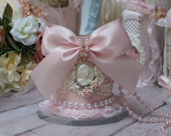 May include: A pink decorative jar with a large pink satin bow, pearl accents, and a cameo image. The jar is surrounded by a pearl and lace trim and a pink pearl necklace.