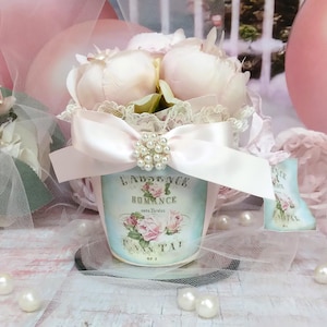 May include: A small, round, light blue container with a floral design and the text "LAURENCE ROMANCE" holds pale pink peonies. A light pink satin bow with a pearl cluster adorns the container. Decorative pearls are scattered around the base.