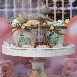 May include: Two decorative Easter pots with bunny and floral designs, filled with colorful eggs and decorative accents. The pots sit on a white, distressed wooden stand, with a wooden bunny figurine in the background. The overall theme is Easter decor.