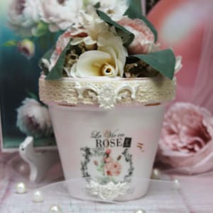 May include: A white ceramic planter with a lace trim and a floral design. The planter has the text "La Vie en Rose" printed on it. The planter is filled with artificial flowers, including pink roses and white hydrangeas.