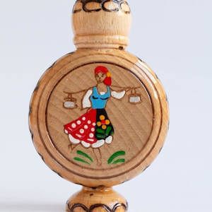 May include: A wooden bottle with a carved design and a painted image of a woman in a red and white dress carrying two buckets. The bottle has a round body and a small, pointed top.