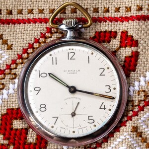 May include: A silver pocket watch with a white face, black hands, and Arabic numerals. The watch is labelled "Kienzle" and "Antimagnetic." It has a gold-coloured loop and is set against a patterned, embroidered fabric with red, white, and brown designs.