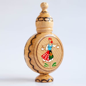 May include: A wooden bottle with a round body and a domed top. The bottle is decorated with a painted image of a woman in a traditional folk costume carrying two buckets.