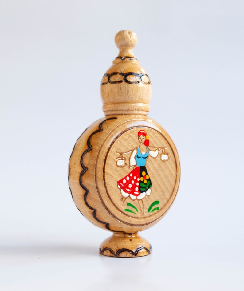 May include: A wooden bottle with a round body and a domed top. The bottle is decorated with a painted image of a woman in a traditional folk costume carrying two buckets.