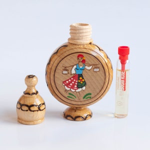 May include: A wooden bottle with a round, painted design of a woman in a traditional folk costume. The bottle has a wooden stopper and is accompanied by a small glass bottle of perfume labeled "Bulgarian Rose".