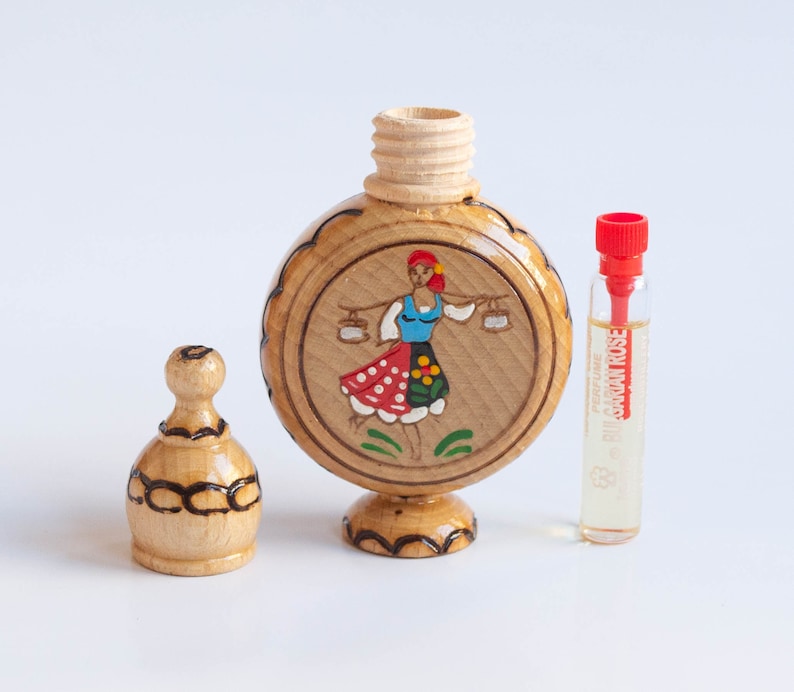 May include: A wooden bottle with a round, painted design of a woman in a traditional folk costume. The bottle has a wooden stopper and is accompanied by a small glass bottle of perfume labeled "Bulgarian Rose".