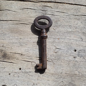May include: A rusty metal key with an oval-shaped head and a long, thin shaft. The key is lying on a weathered wooden surface.