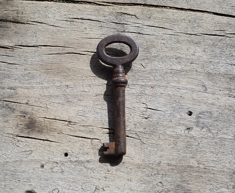 May include: A rusty metal key with an oval-shaped head and a long, thin shaft. The key is lying on a weathered wooden surface.