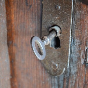 May include: A close-up of a rusty key inserted into a keyhole on a wooden door. The keyhole is surrounded by a metal plate.