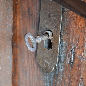 May include: A close-up of an antique key inserted into a rusty metal lock on a wooden door. The key is made of metal and has a simple design. The lock is attached to the door with screws.