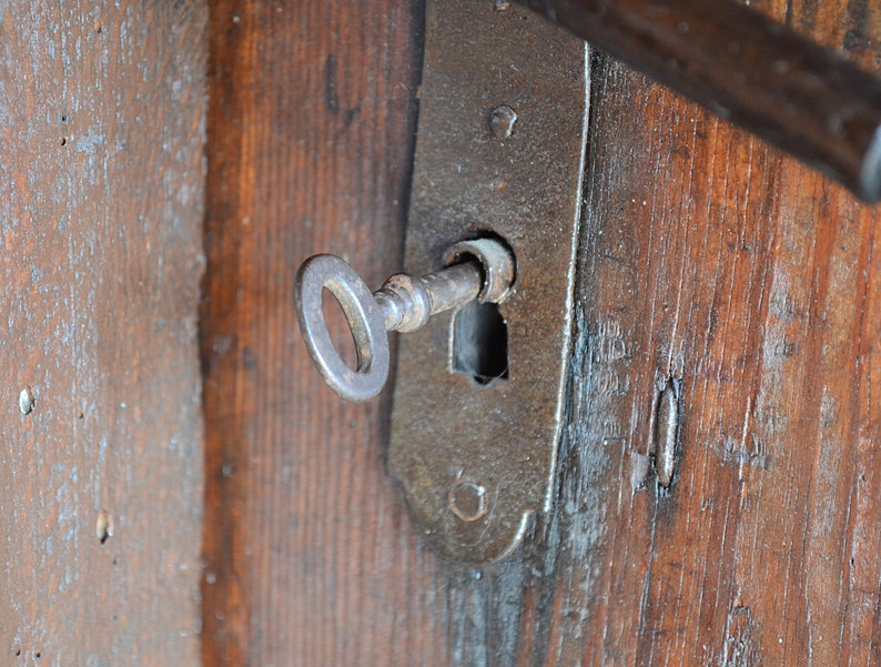 May include: A close-up of an antique key inserted into a rusty metal lock on a wooden door. The key is made of metal and has a simple design. The lock is attached to the door with screws.