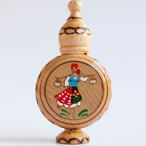 May include: A wooden bottle with a round body and a domed top. The bottle is decorated with a painted image of a woman in a red, white, and blue dress carrying two buckets.