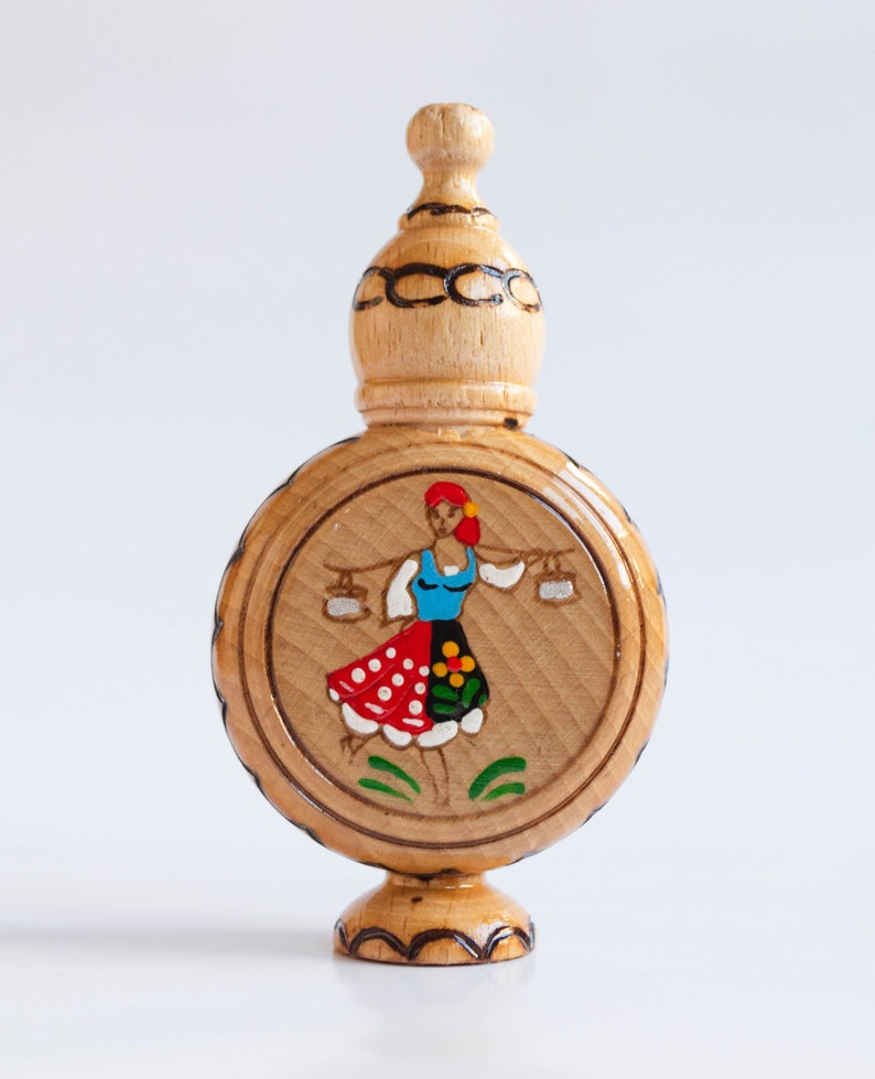 May include: A wooden bottle with a round body and a domed top. The bottle is decorated with a painted image of a woman in a red, white, and blue dress carrying two buckets.