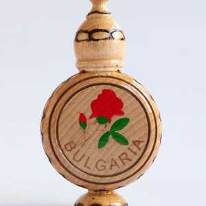 May include: A wooden bottle with a round, carved base and a domed top. The bottle is decorated with a red rose and green leaves, and the word "BULGARIA" is carved around the rose.