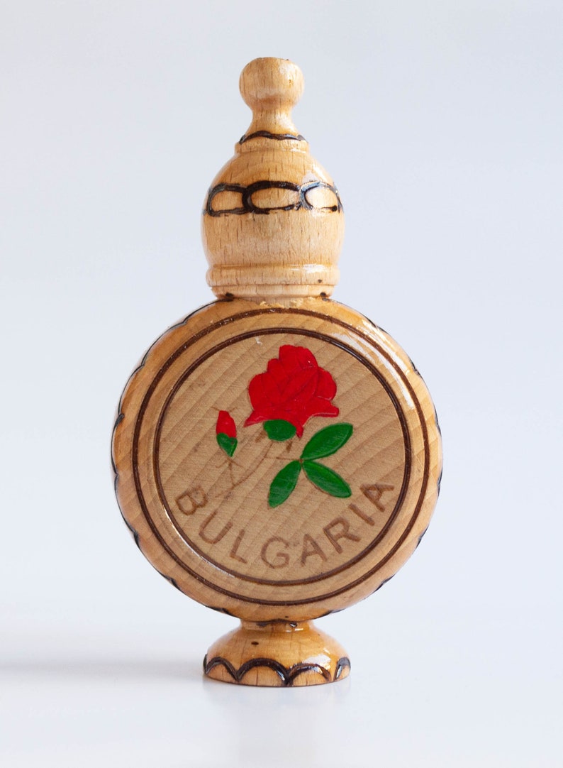 May include: A wooden bottle with a round, carved base and a domed top. The bottle is decorated with a red rose and green leaves, and the word "BULGARIA" is carved around the rose.