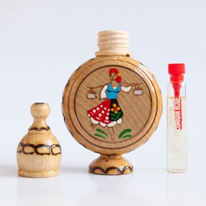 May include: A wooden bottle with a hand-painted design of a woman in a red and white dress holding two buckets. The bottle has a wooden stopper and sits on a wooden base. A small glass bottle of perfume labeled "Bulgarian Rose" is next to the wooden bottle.