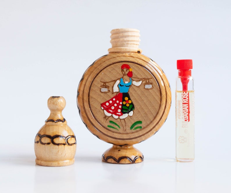 May include: A wooden bottle with a hand-painted design of a woman in a red and white dress holding two buckets. The bottle has a wooden stopper and sits on a wooden base. A small glass bottle of perfume labeled "Bulgarian Rose" is next to the wooden bottle.