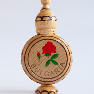 May include: A wooden bottle with a carved top and a round base. The bottle is painted with a red rose and green leaves. The word "BULGARIA" is written around the rose.