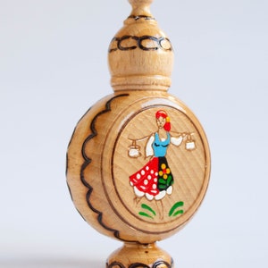 May include: A wooden bottle with a carved design and a painted image of a woman in a traditional folk costume. The bottle has a round body and a small, pointed top.