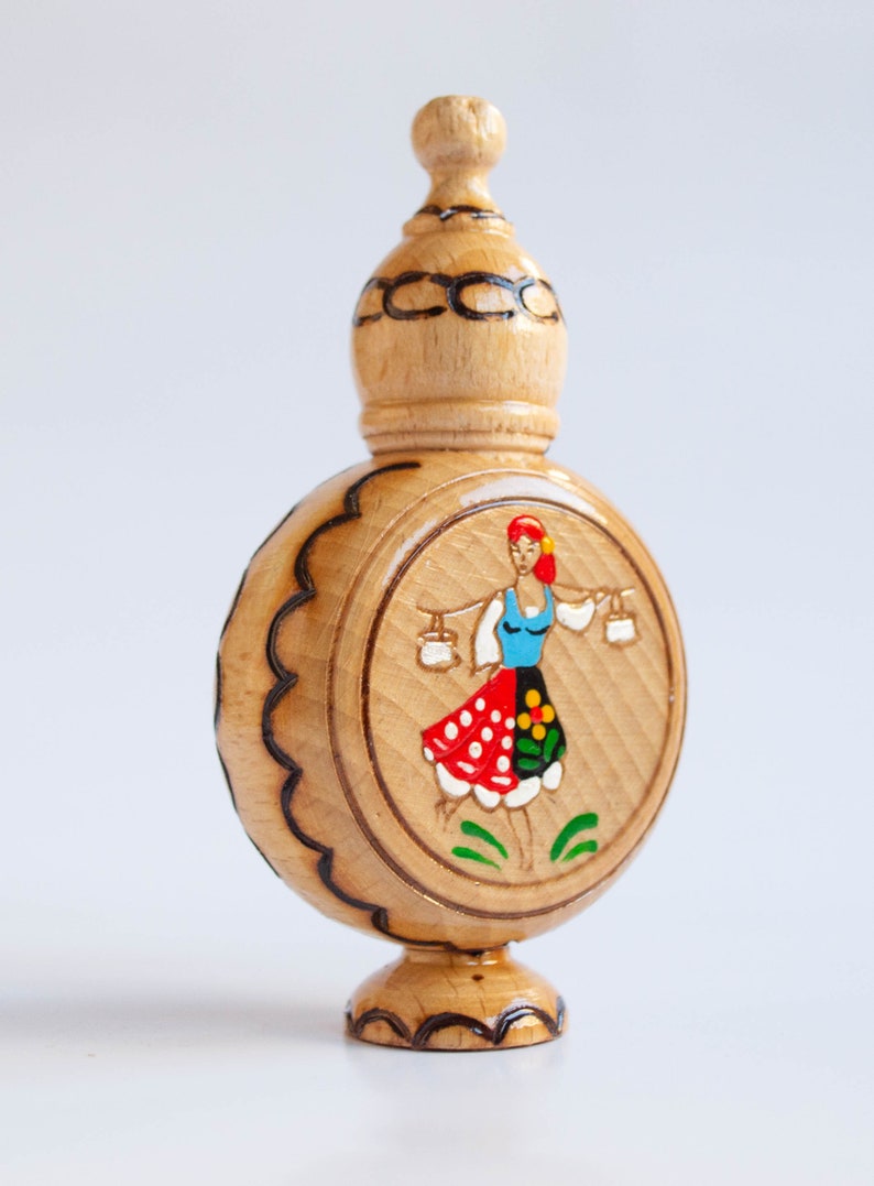 May include: A wooden bottle with a carved design and a painted image of a woman in a traditional folk costume. The bottle has a round body and a small, pointed top.