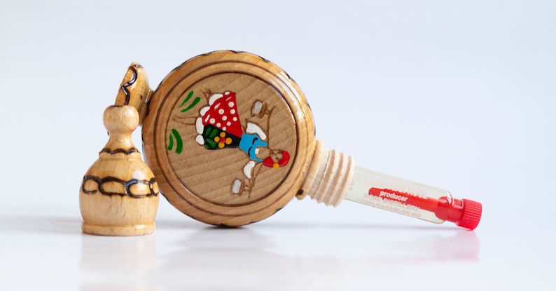 May include: A wooden bottle with a painted design of a woman in a red and white dress. The bottle has a cork stopper and a small glass vial attached to it. The vial is labeled "producer".