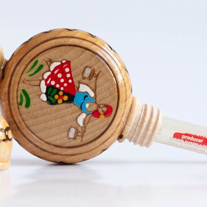 May include: A wooden bottle with a painted design of a woman in a red and blue dress. The bottle has a cork stopper and a small glass vial labeled "producer".