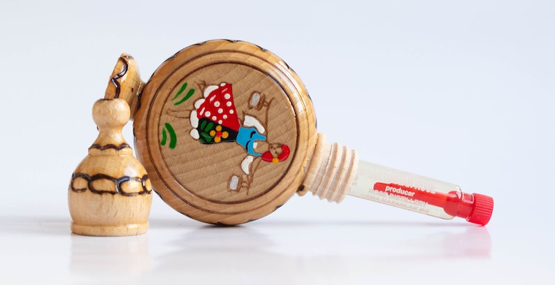 May include: A wooden bottle with a painted design of a woman in a red and blue dress. The bottle has a cork stopper and a small glass vial labeled "producer".