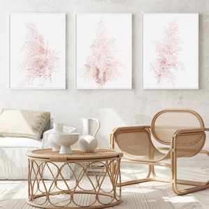 Puede incluir: Tres impresiones enmarcadas de hierba de la pampa rosa sobre un fondo blanco. Las impresiones est&aacute;n colgadas en una pared blanca sobre una silla de mimbre y una mesa de caf&eacute; redonda de mimbre.