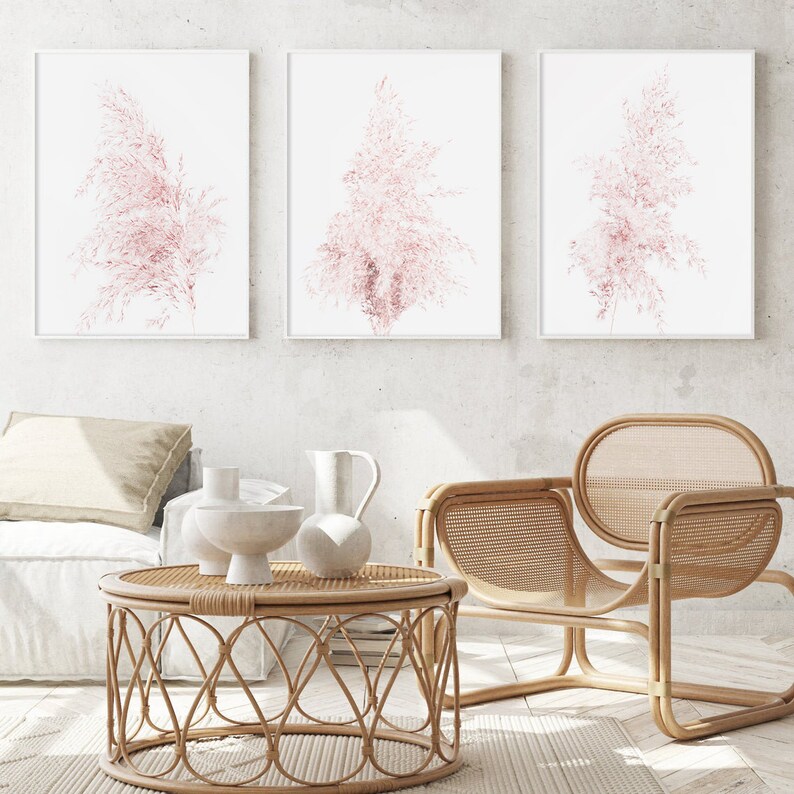 Puede incluir: Tres impresiones enmarcadas de hierba de la pampa rosa sobre un fondo blanco. Las impresiones est&aacute;n colgadas en una pared blanca sobre una silla de mimbre y una mesa de caf&eacute; redonda de mimbre.