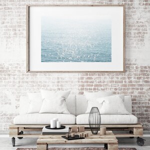 May include: A white couch with two white pillows sits in front of a brick wall. A framed photograph of a sparkling blue ocean hangs above the couch. A wooden coffee table with a black metal vase sits in front of the couch.